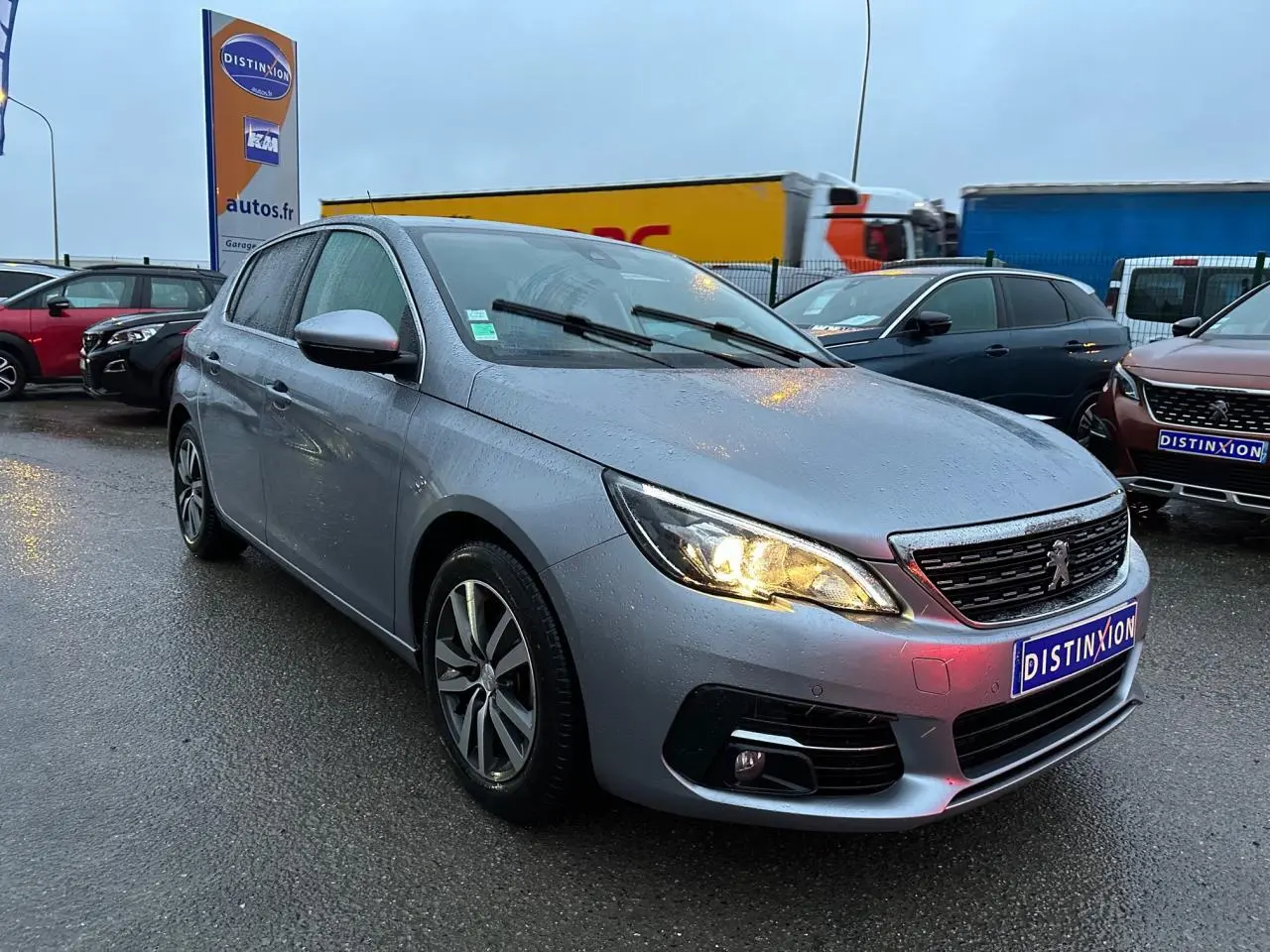 PEUGEOT 308 gris Artense en 3/4 avant droit sous la pluie, phare allumé et jantes alliage visibles.