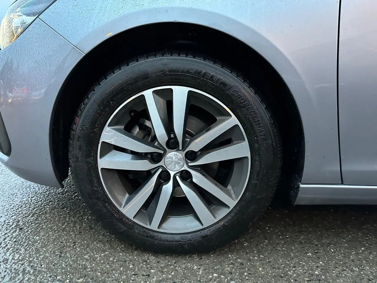 Gros plan sur la roue avant gauche d'une Peugeot 308 gris Artense avec jante alliage et carrosserie humide.