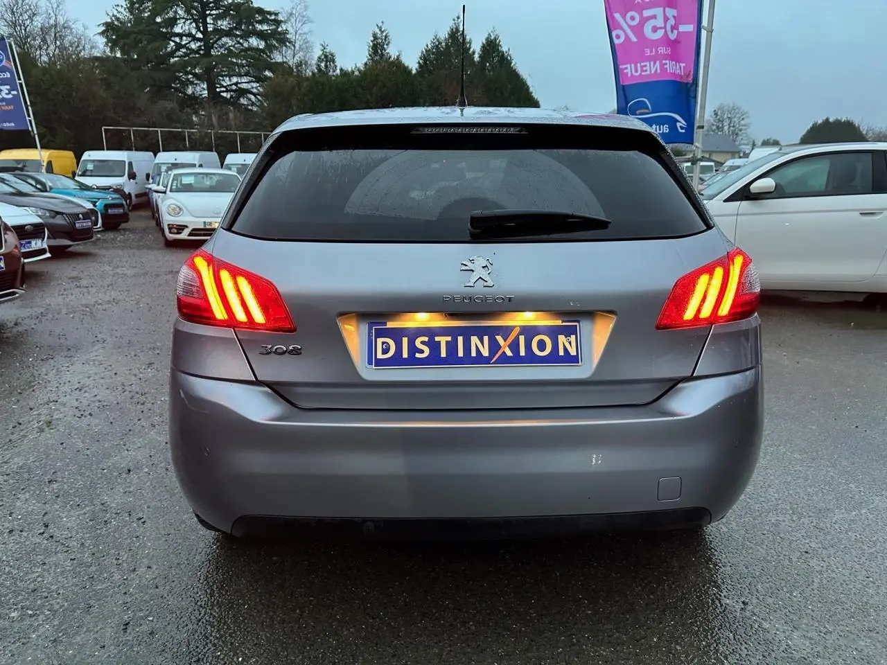 Vue arrière d'une Peugeot 308 gris Artense avec feux arrière allumés sur un parking humide.
