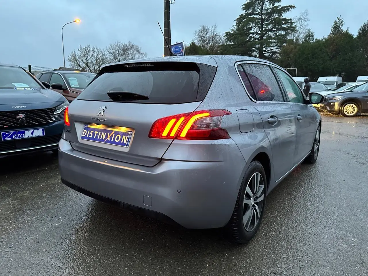 Peugeot 308 gris Artense vue 3/4 arrière droit avec feux arrière allumés sur parking humide.