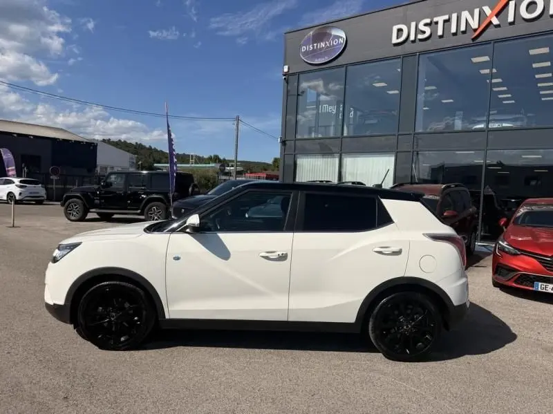 Vue de profil côté gauche d'un SsangYong Tivoli blanc avec toit noir et jantes noires, devant un bâtiment commercial.