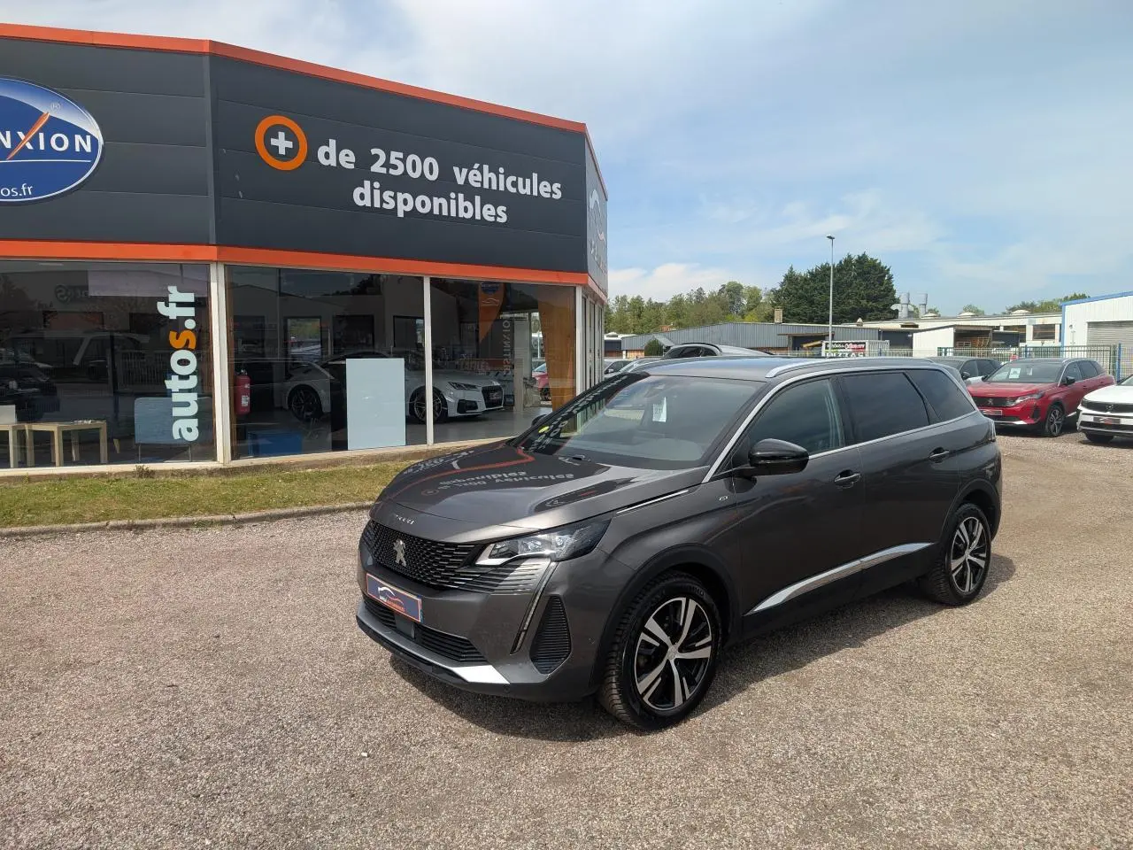 Peugeot 5008 gris métallisé vue 3/4 avant droit avec jantes bi-ton et calandre GT devant un concessionnaire.