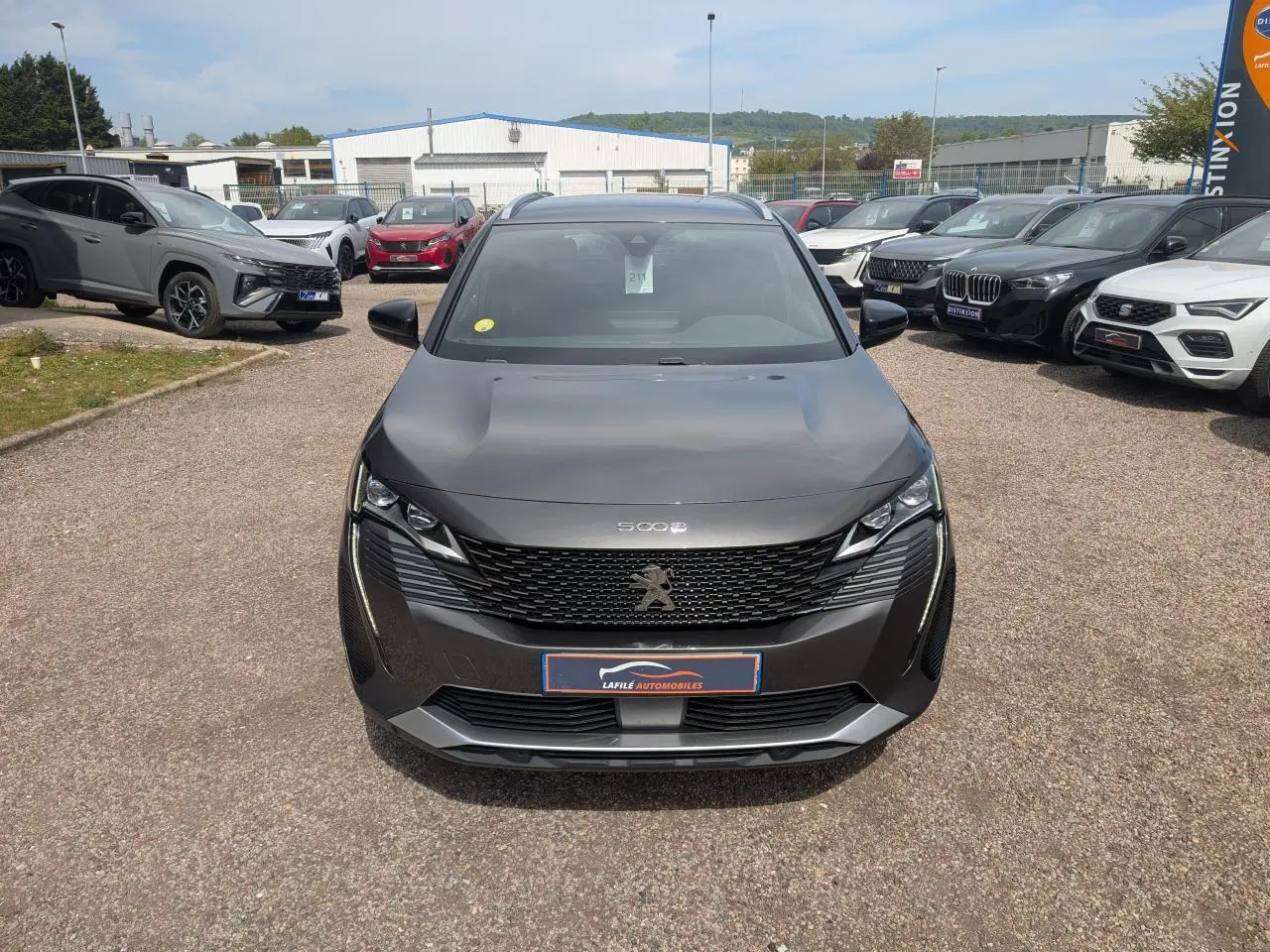 Peugeot 5008 gris métallisé vue de face, avec calandre GT noire et feux LED 3D, sur parking extérieur.