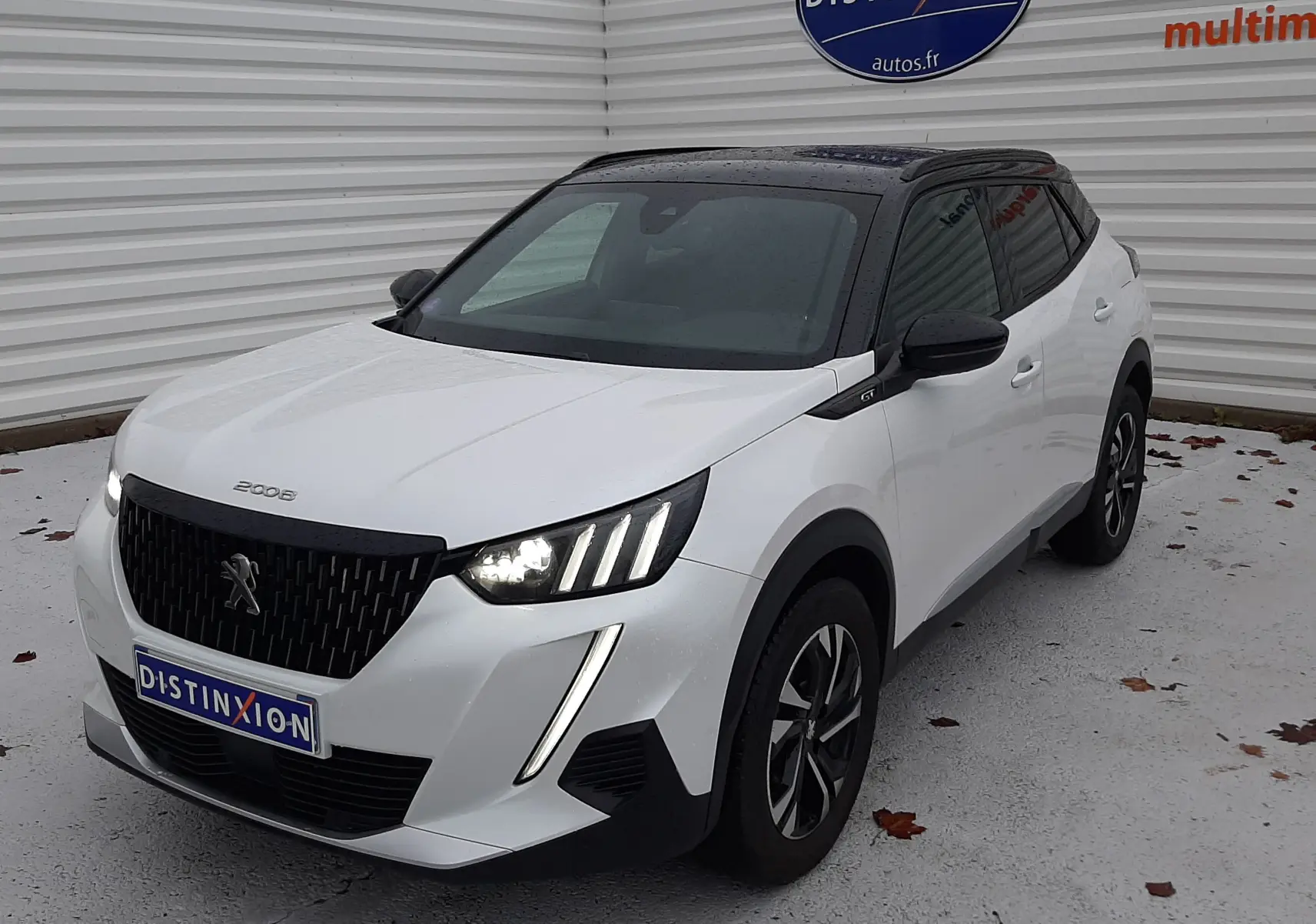 Peugeot 2008 blanc nacré avec toit noir en 3/4 avant droit, phares allumés et attelage visible à l'arrière.