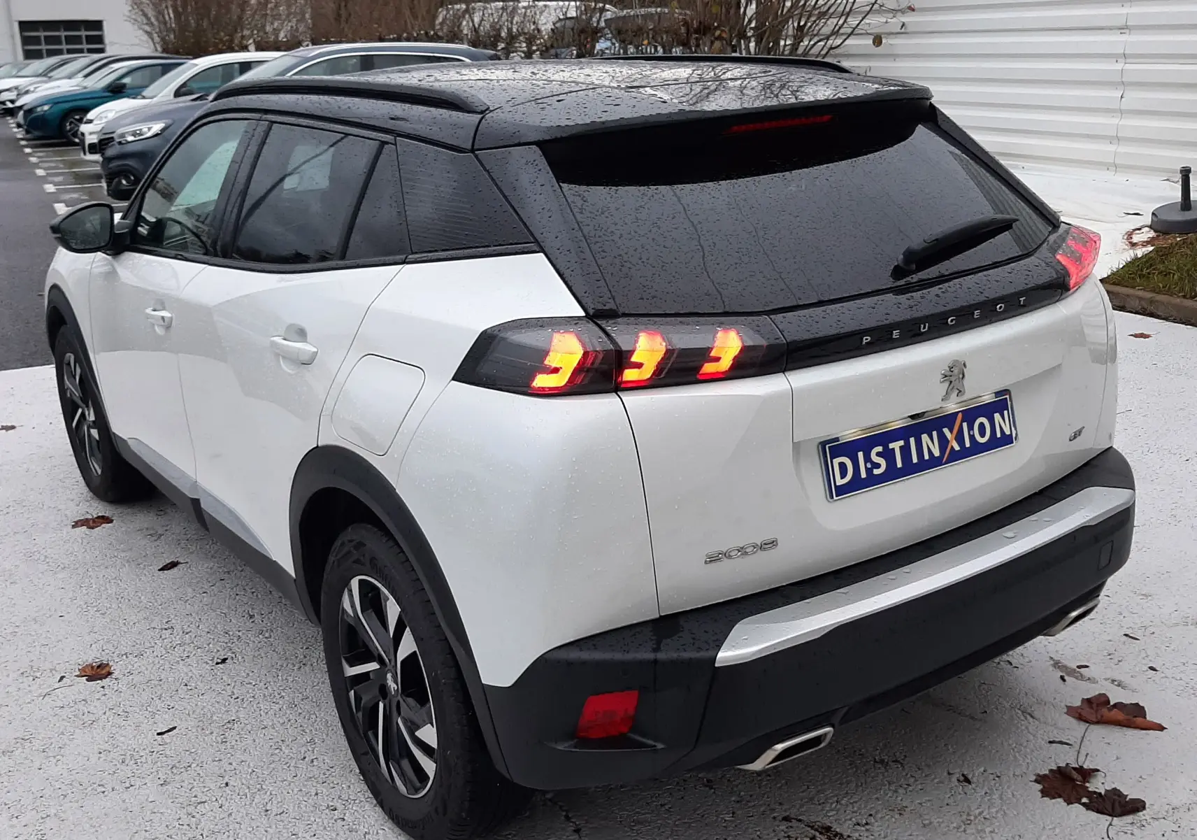 Vue 3/4 arrière droite d'un Peugeot 2008 blanc nacré avec toit noir et feux arrière allumés sur parking extérieur.