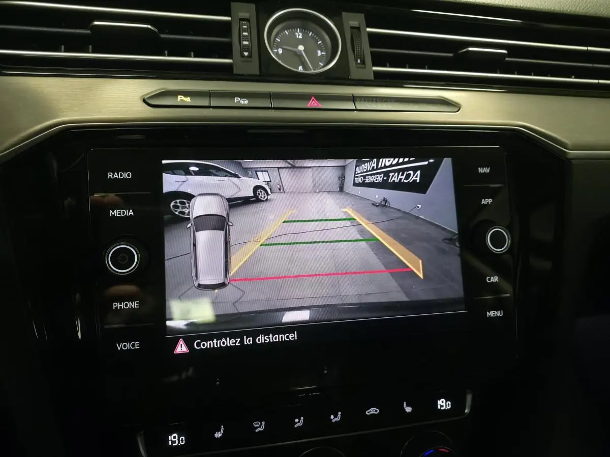Écran tactile de la Volkswagen Passat SW gris montrant la caméra de recul avec lignes de guidage colorées et horloge analogique au-dessus.