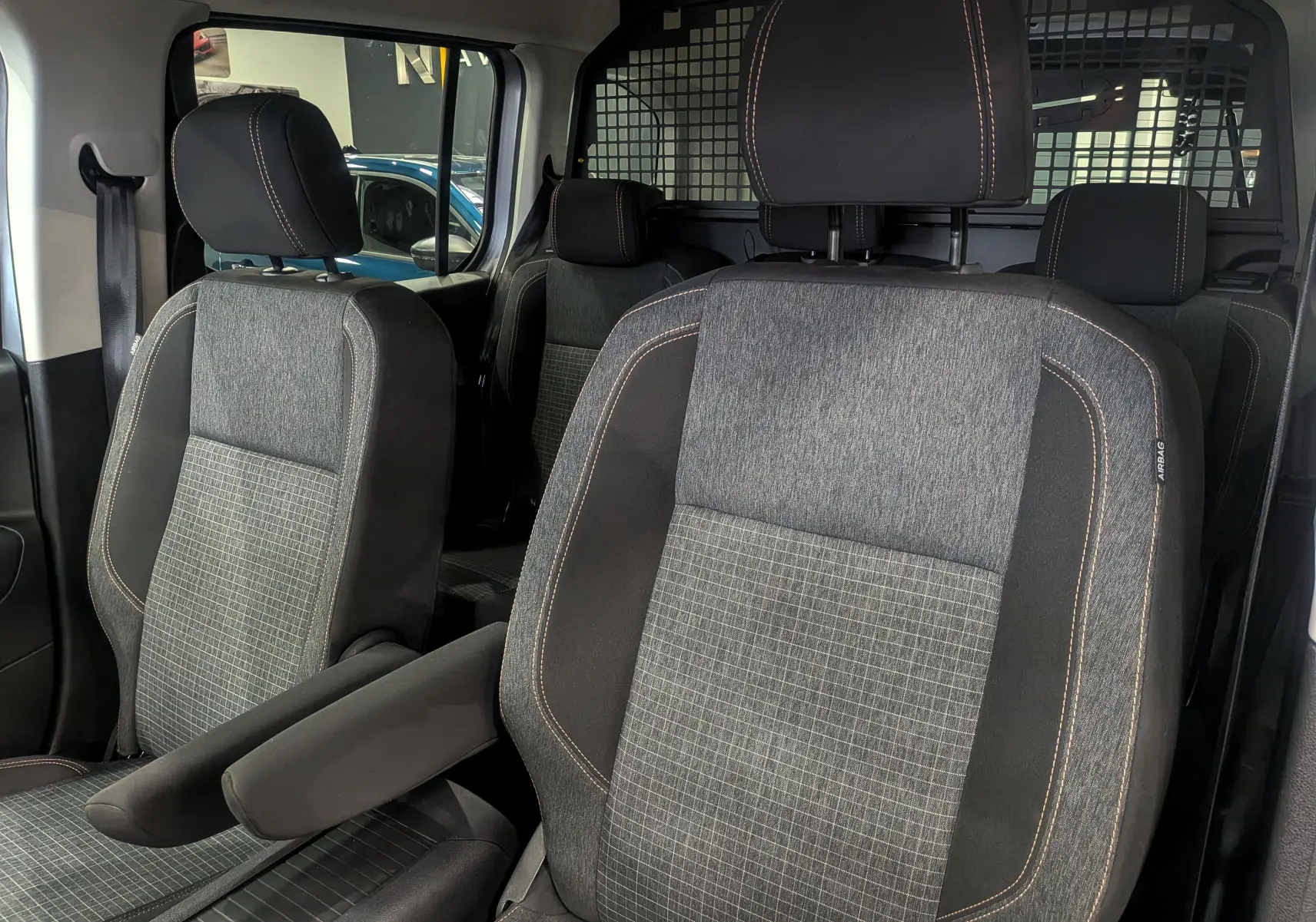 Gros plan sur les sièges avant gris à surpiqûres orange du Peugeot Rifter 2024, avec séparation grillagée arrière visible.