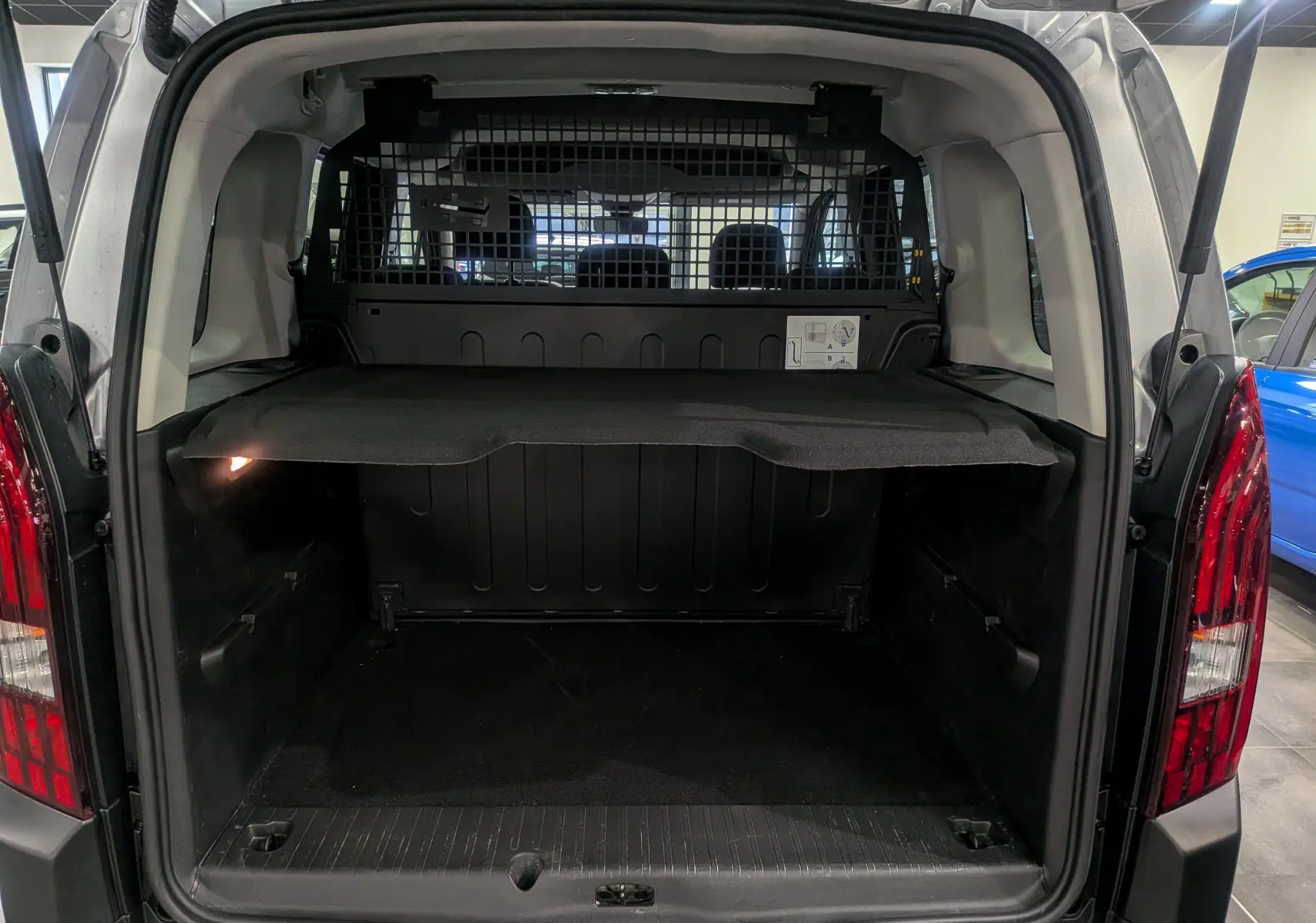 Coffre ouvert vu de l’arrière d’un Peugeot Rifter gris Artense 2024 avec séparation grillagée et tablette de rangement.