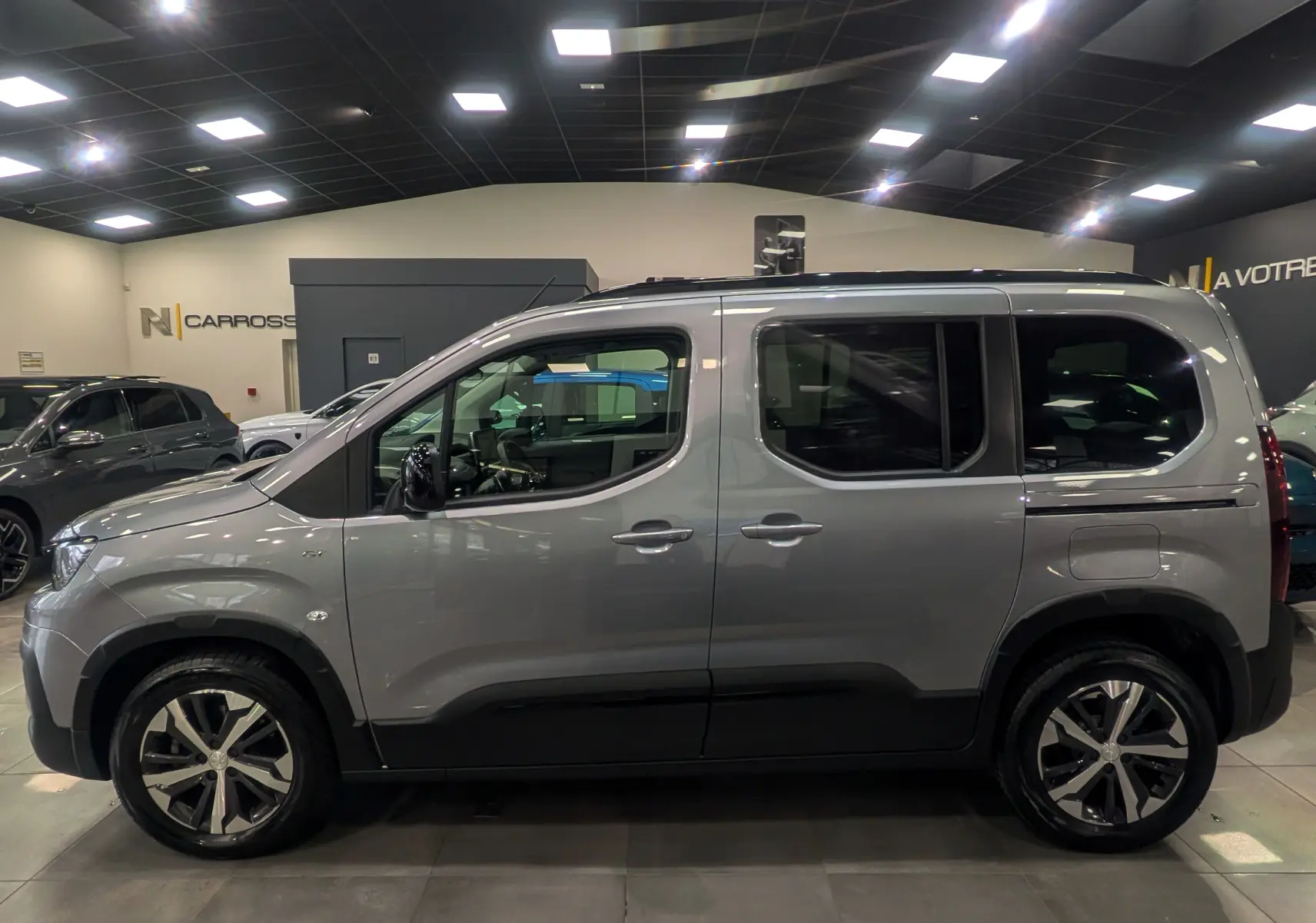 Peugeot Rifter 2024 gris Artense vu de profil côté gauche en intérieur, avec jantes bi-ton et toit noir.