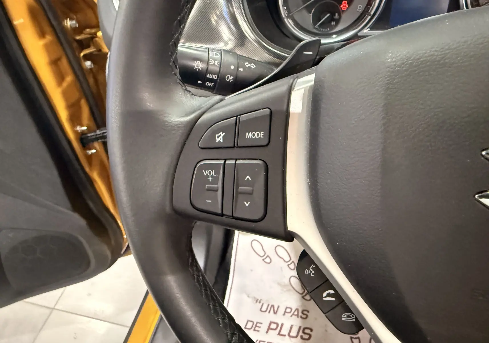 Gros plan sur le volant cuir noir du Suzuki Vitara jaune toit noir, avec commandes audio et porte conducteur ouvert.