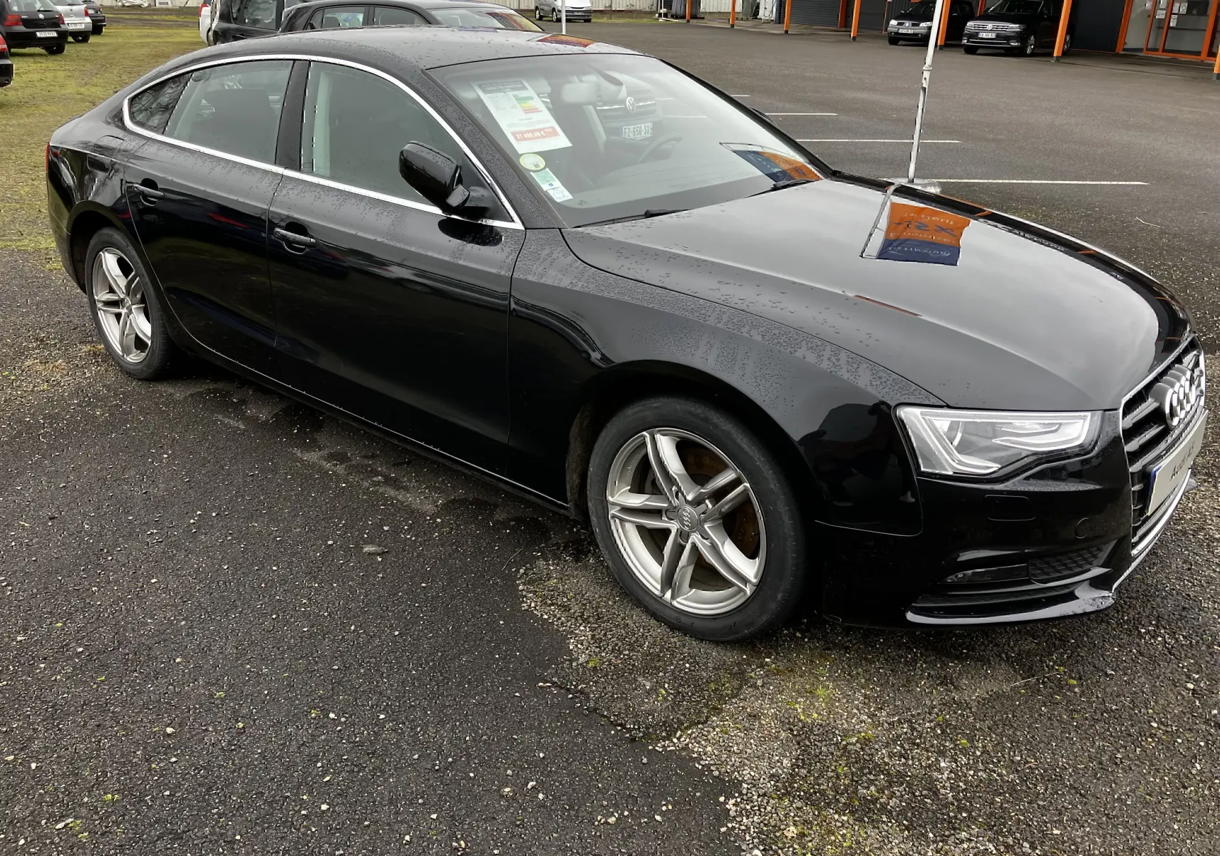 Audi A5 Sportback noir vue 3/4 avant côté gauche, avec jantes alliage et carrosserie brillante sous la pluie.