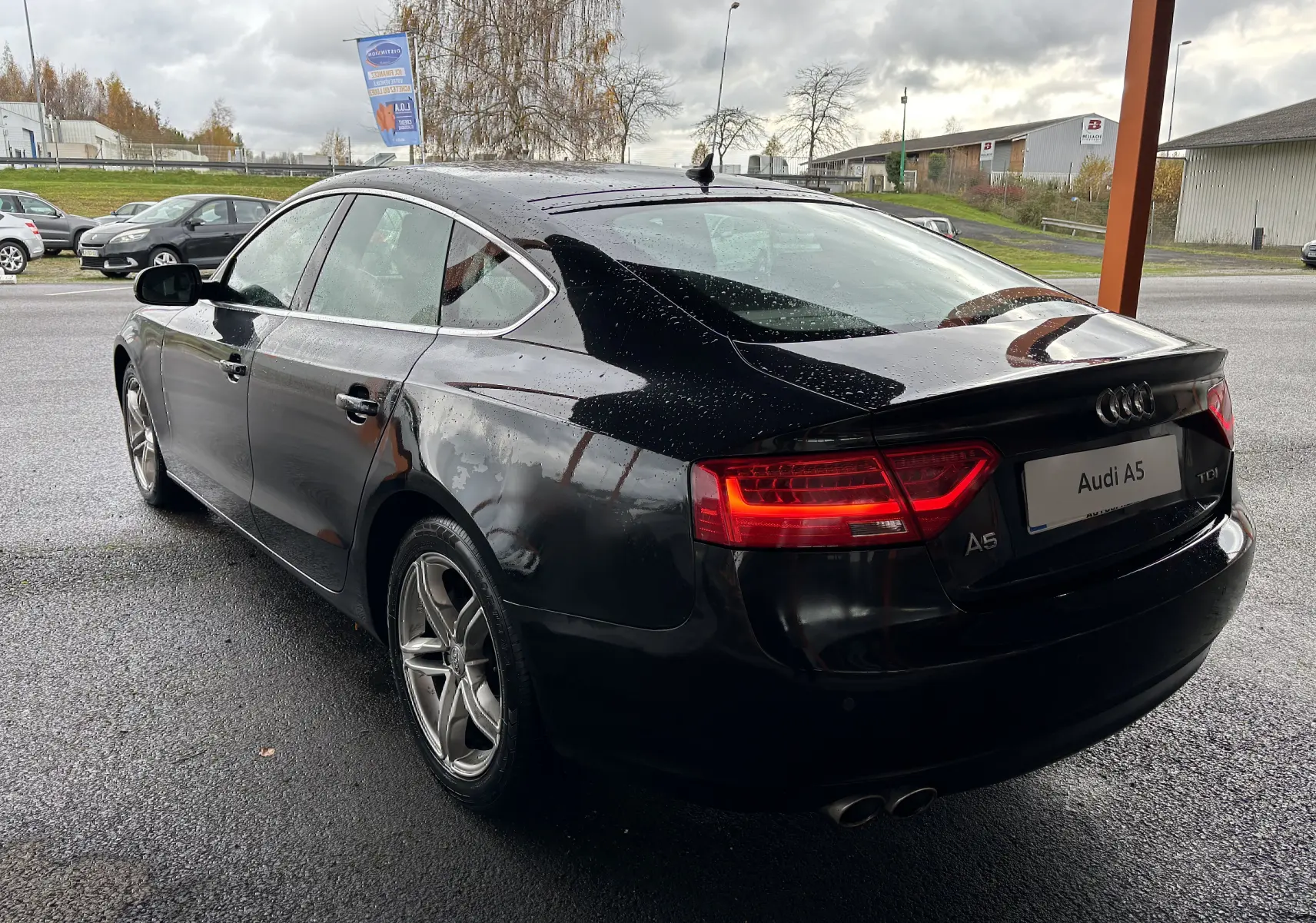 Audi A5 Sportback noir vue 3/4 arrière droit sous la pluie avec feux arrière allumés et jantes alliage.