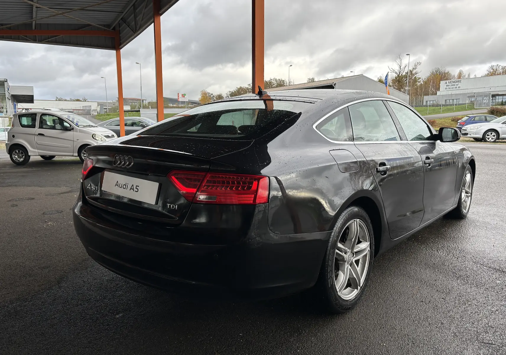Audi A5 Sportback noir vue 3/4 arrière droit sous abri, avec feux arrière allumés et jantes alliage cinq branches.