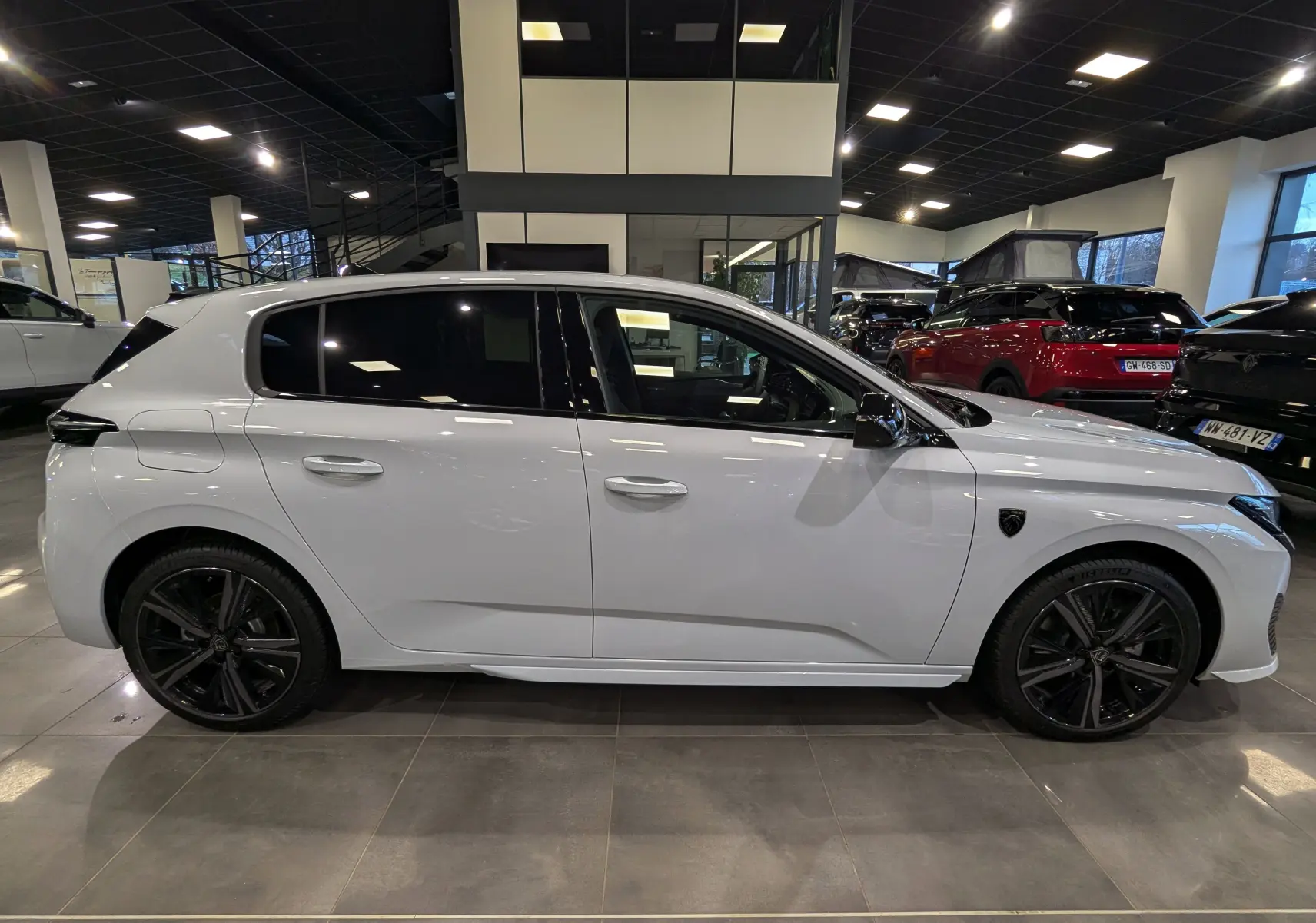 Profil côté gauche d'une Peugeot 308 GT blanche Okénite avec jantes noires et vitres teintées en showroom.