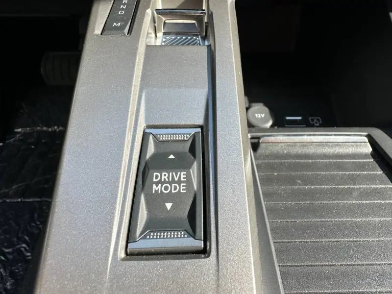 Gros plan sur la console centrale grise de la Peugeot 308 2025 avec bouton Drive Mode et prises USB et 12V visibles.