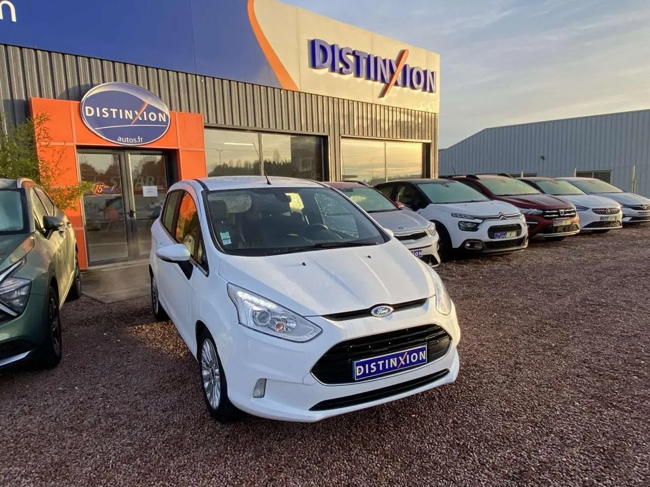 Ford B-MAX blanc vue 3/4 avant droit sur un parking devant un concessionnaire Distinxion au coucher du soleil.