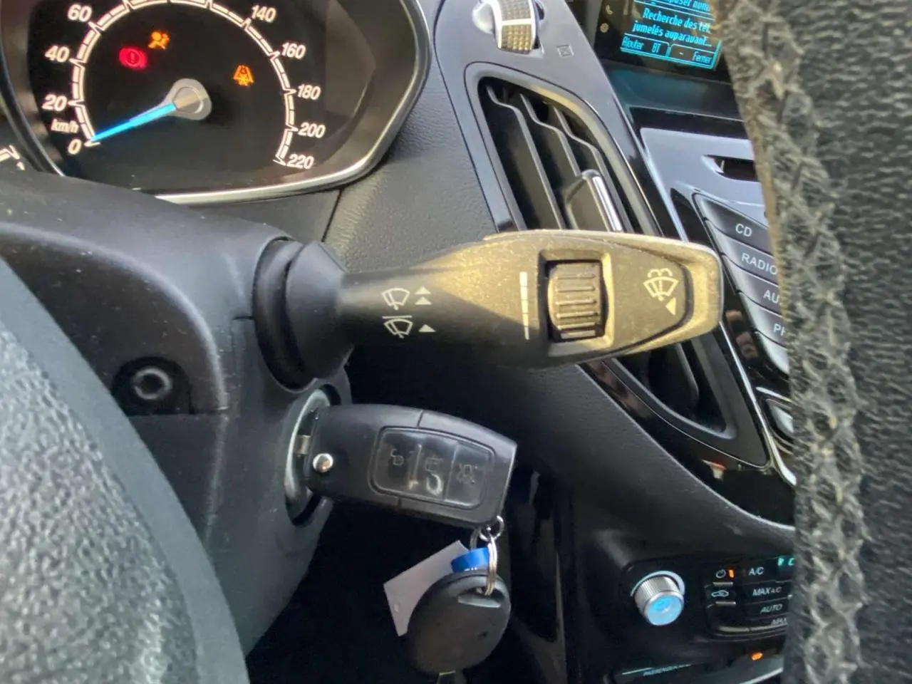 Gros plan sur la colonne de direction et le tableau de bord d’une Ford B-MAX blanche de 2014, avec clé insérée et commandes d’essuie-glace visibles.