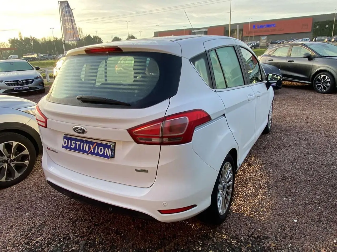 Ford B-MAX blanc en 3/4 arrière droit sur parking, avec jantes alliage et feux arrière distinctifs.