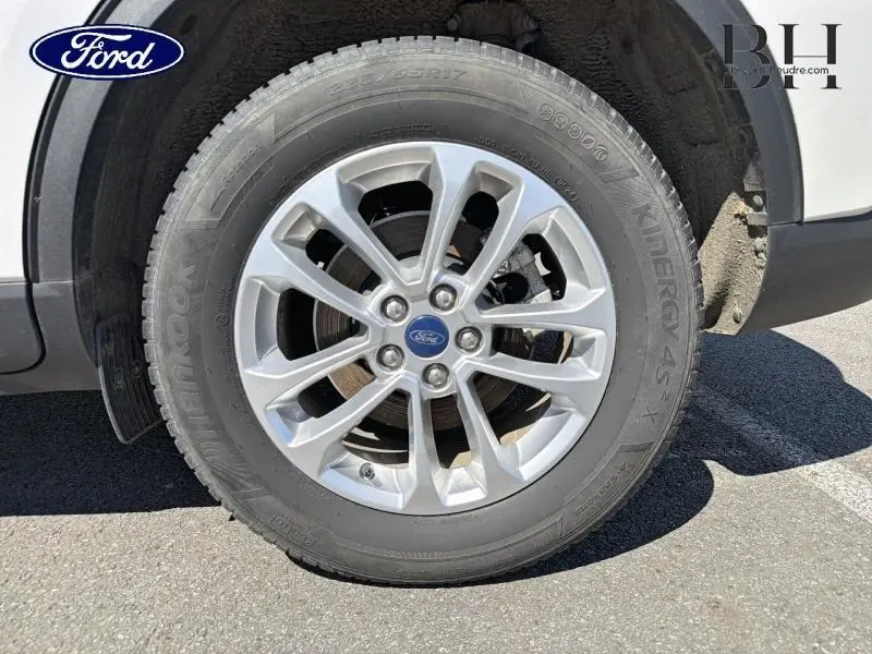 Gros plan sur la roue avant gauche du Ford Kuga blanc Glacier, avec jante alliage et logo Ford central.