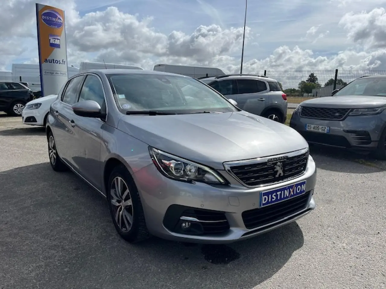 Vue 3/4 avant d'une Peugeot 308 gris clair métallisé garée sur un parking extérieur sous ciel partiellement nuageux.