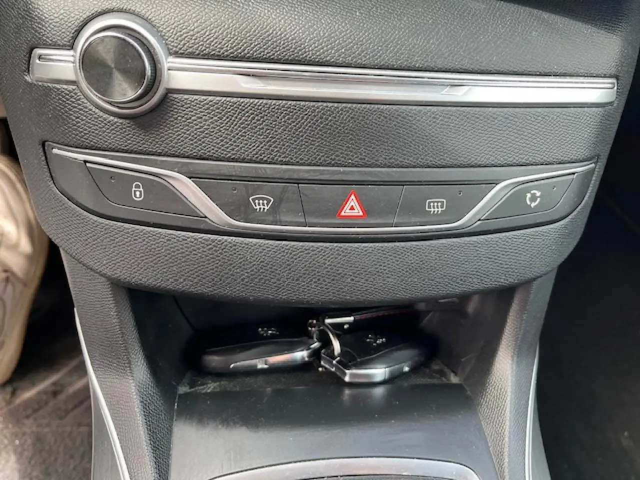 Gros plan sur la console centrale noire de la Peugeot 308 avec les boutons de verrouillage, désembuage et feux de détresse, clés posées dessous.