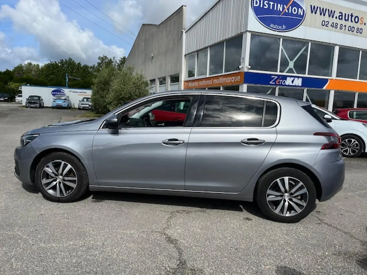 Profil droit d'une Peugeot 308 gris clair métallisé garée devant un bâtiment commercial, avec jantes alliage visibles.