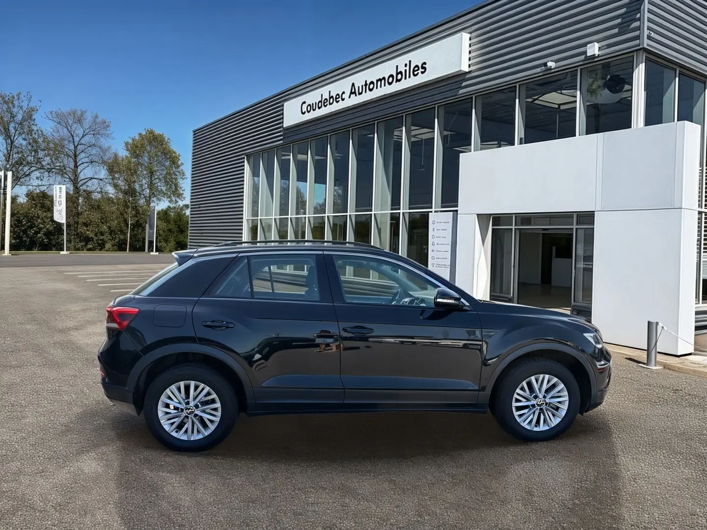 Profil côté gauche du Volkswagen T-Roc noir 2025, avec jantes alliage 16'' et rétroviseurs électriques devant un concessionnaire.
