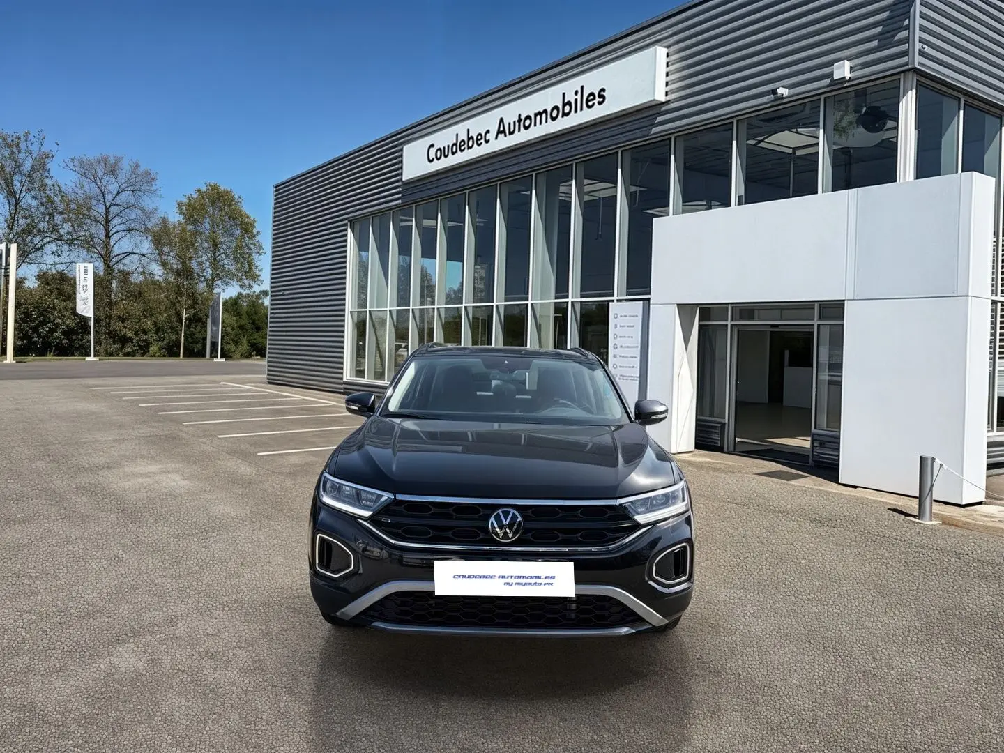 Vue frontale d'un Volkswagen T-Roc noir 2025 garé devant un bâtiment moderne de concession automobile.