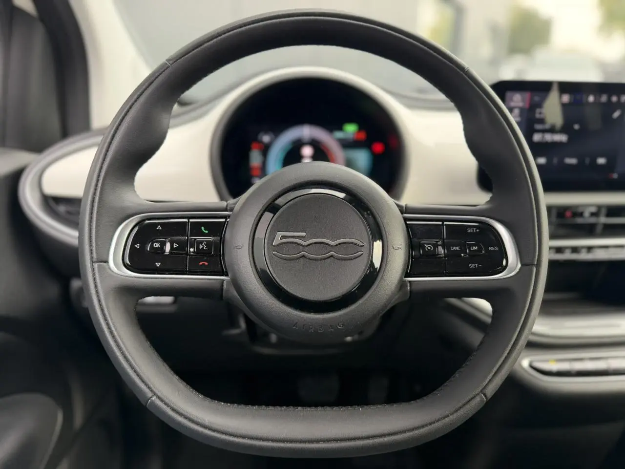 Vue rapprochée du volant noir de la Fiat 500 électrique 2024 avec commandes intégrées et tableau de bord numérique flou.