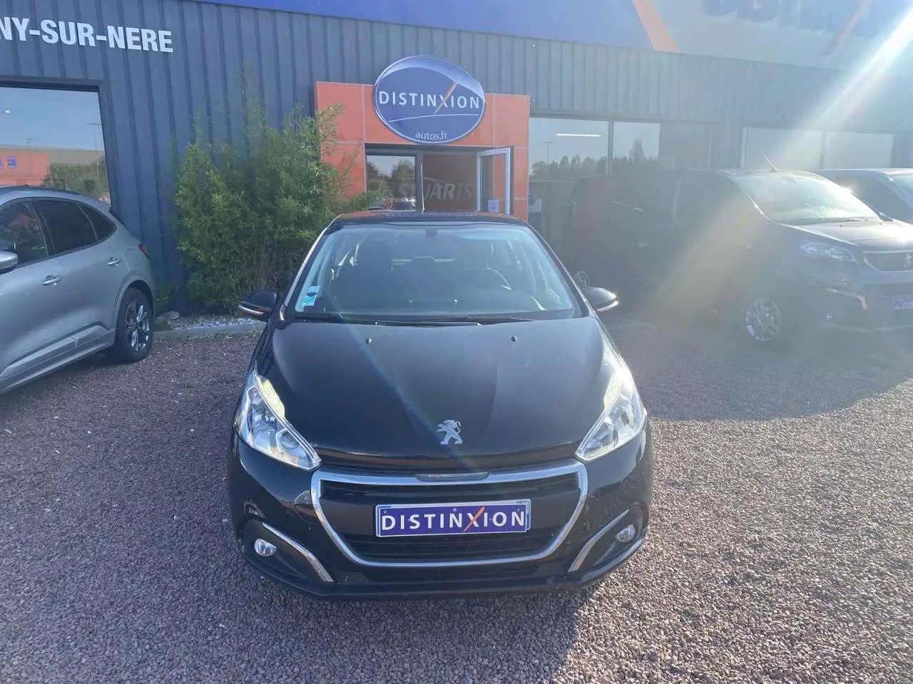 Peugeot 208 noire vue de face, avec calandre chromée et phares allumés, stationnée devant un garage.