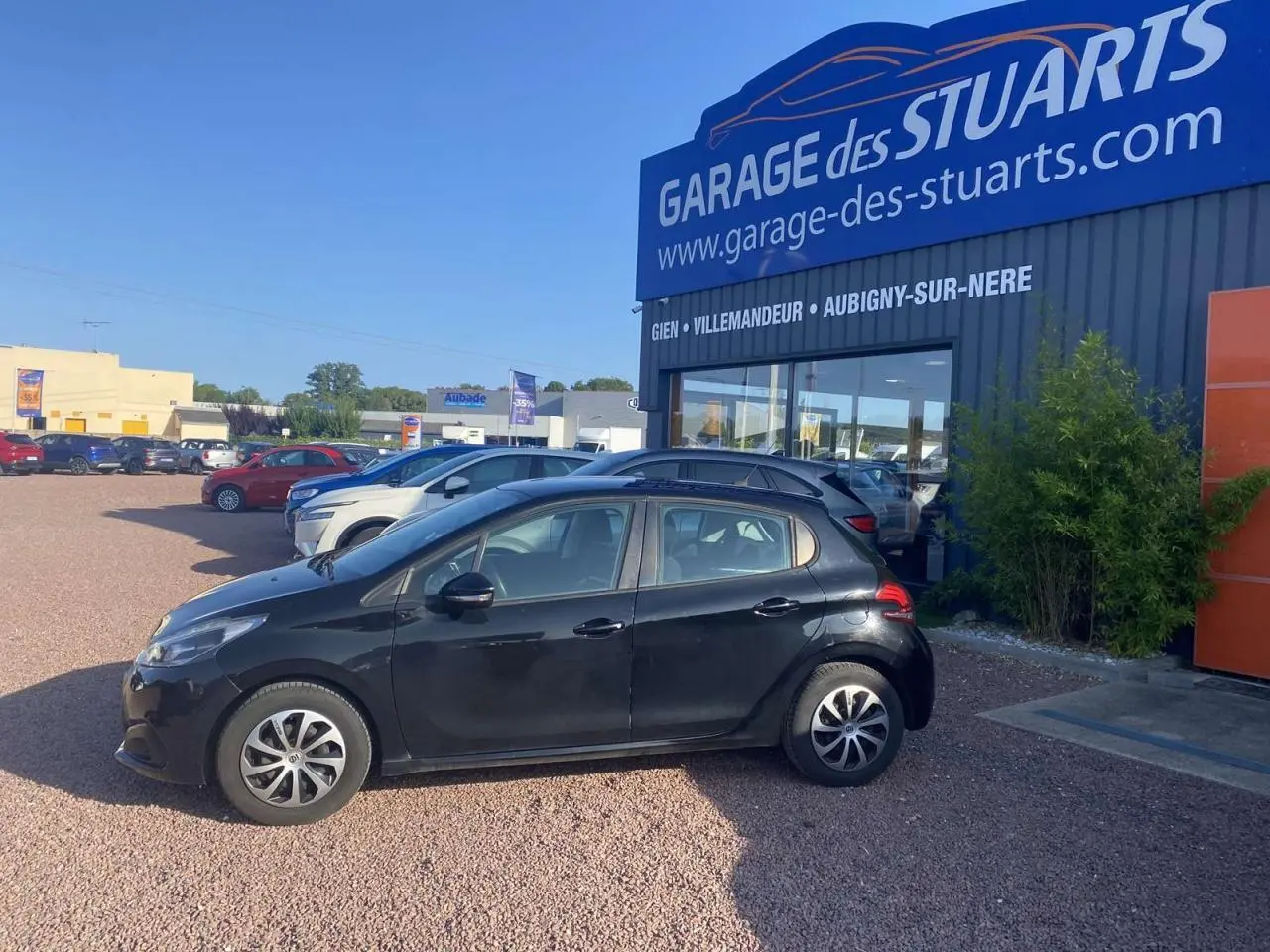 Peugeot 208 noire vue de profil côté gauche, avec jantes acier 15 pouces et roue de secours, stationnée devant un garage.