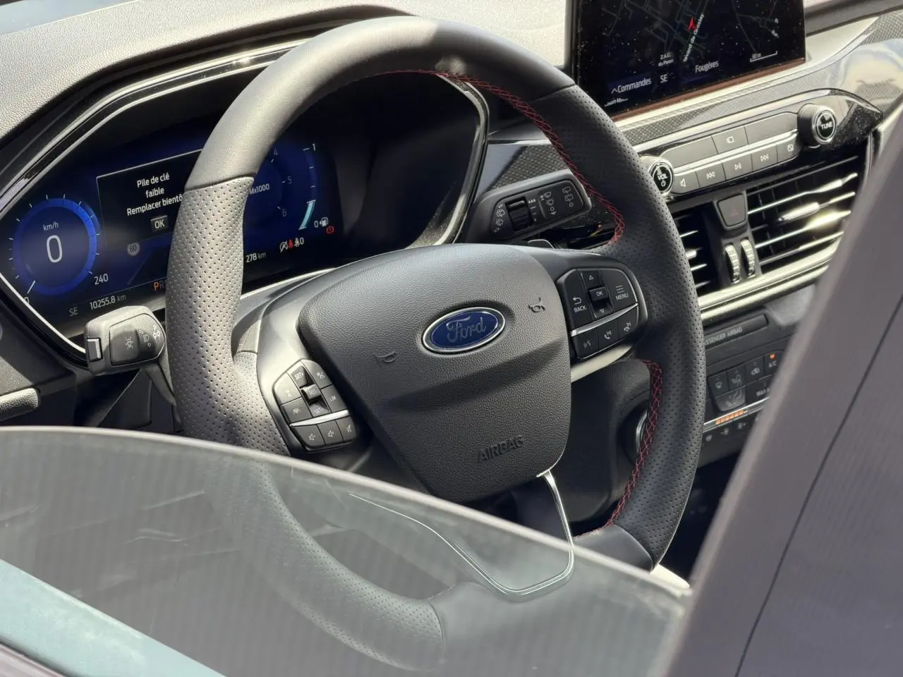 Vue intérieure centrée sur le volant noir perforé avec surpiqûres rouges du Ford Kuga 2023 gris Solar, tableau de bord numérique visible.