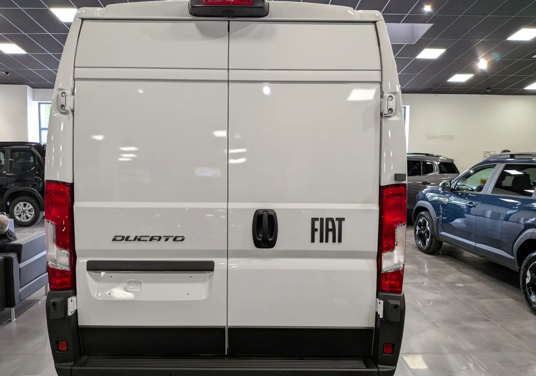 Vue arrière du fourgon blanc Fiat Ducato avec portes battantes et feux arrière rouges en intérieur showroom.