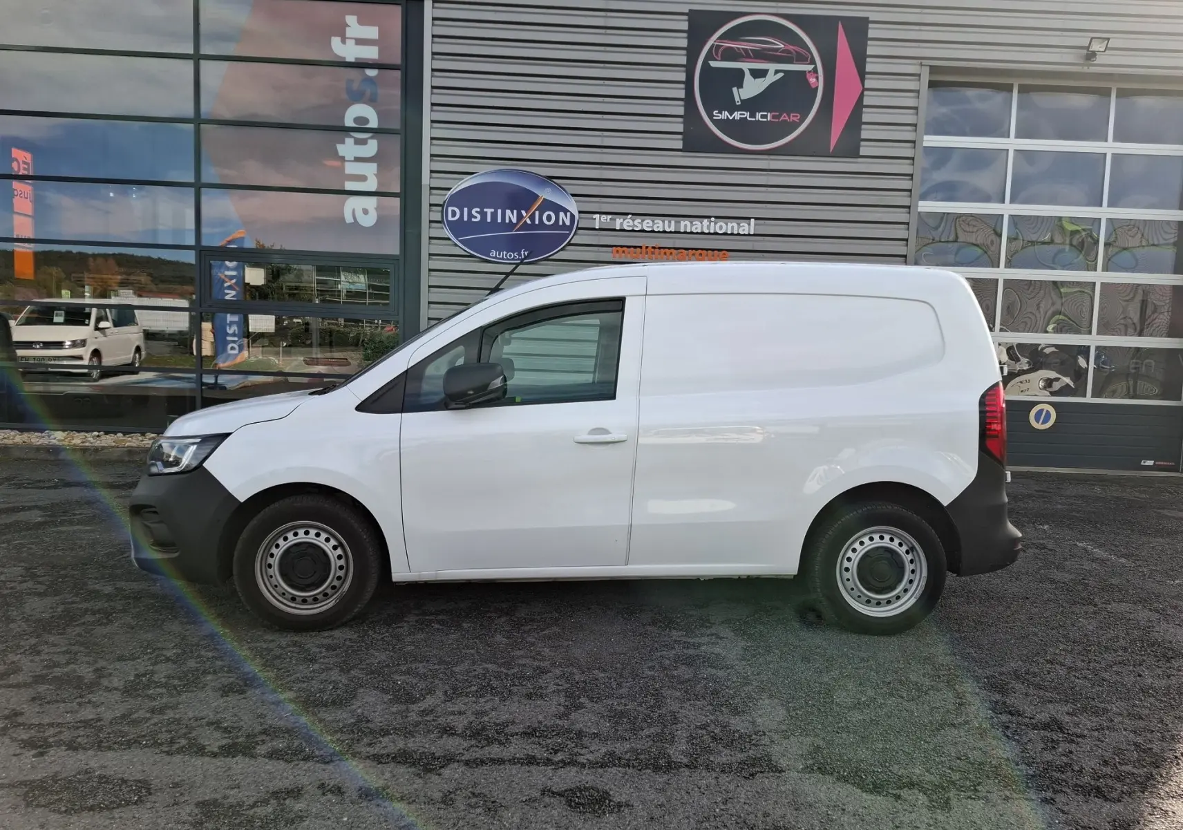 Profil latéral droit d’un Renault Kangoo Pro blanc stationné devant un bâtiment commercial avec jantes acier noires.