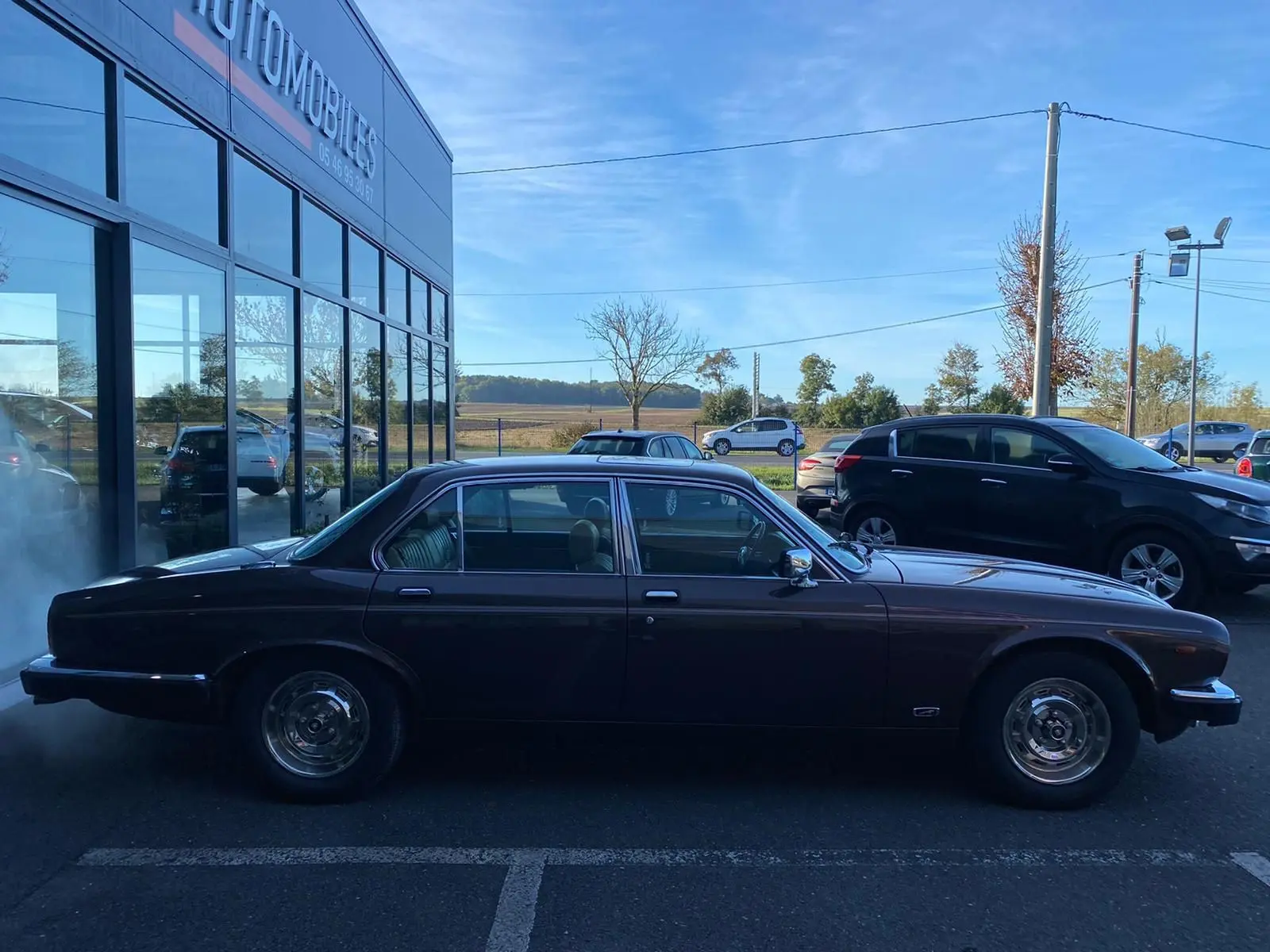 Profil côté gauche d'une Jaguar XJ6 Série 3 4.2 Sovereign marron de 1985 garée devant un bâtiment vitré.