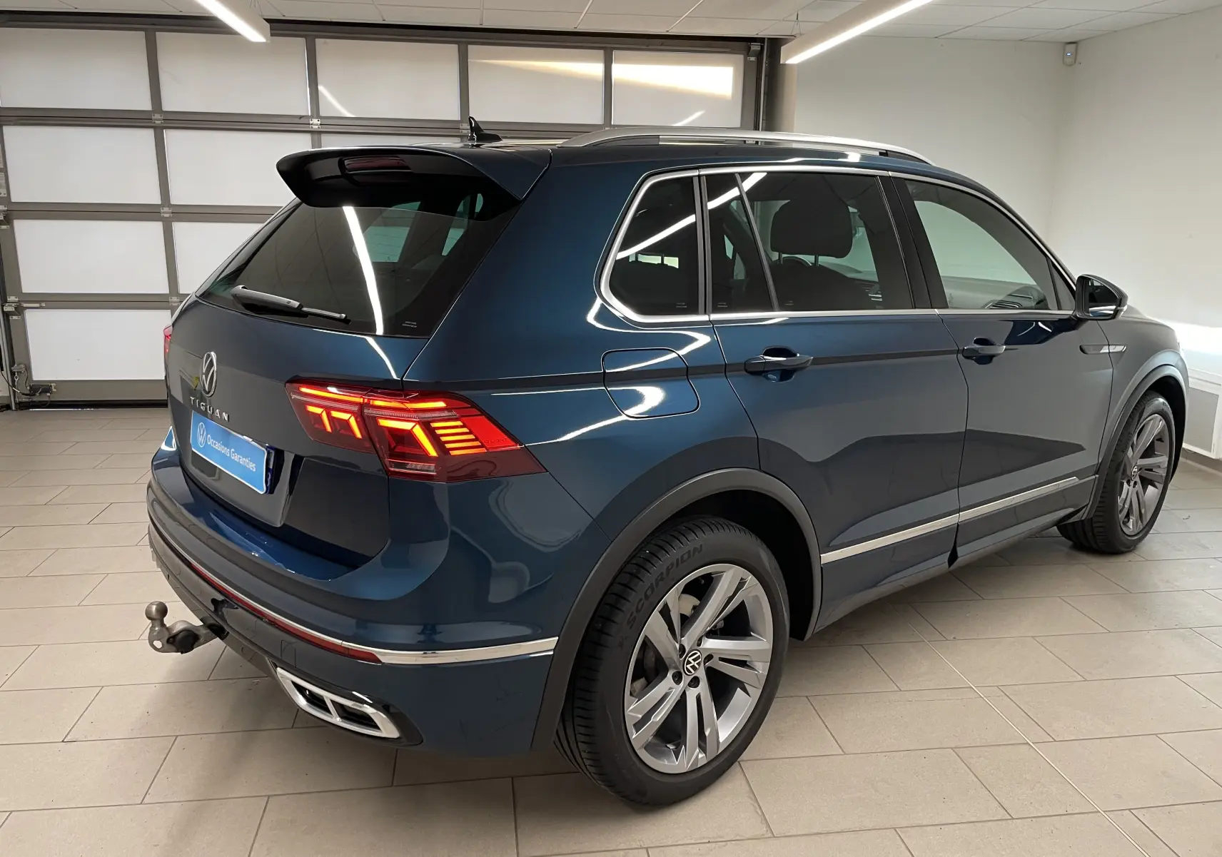 Vue 3/4 arrière droite d'un Volkswagen Tiguan 2024 bleu Nightshade avec attelage escamotable et feux arrière allumés.