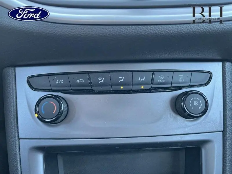 Panneau de commande de climatisation gris dans une Ford, avec boutons et deux molettes rotatives visibles de face.