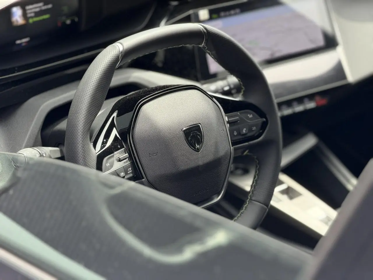 Gros plan sur le volant compact cuir noir avec logo Peugeot, tableau de bord numérique et écran tactile en arrière-plan.