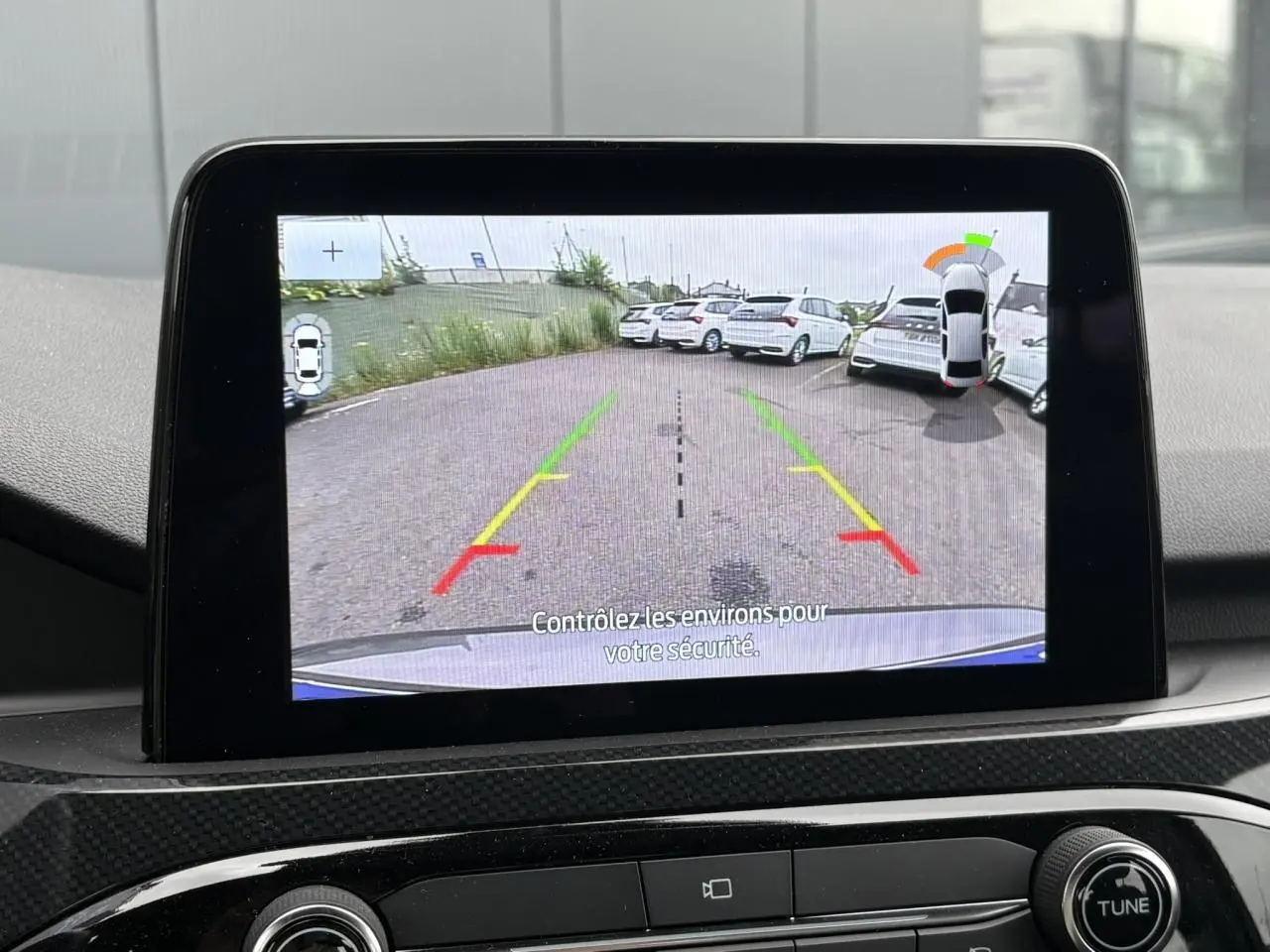Écran de caméra de recul montrant l’arrière d’un Ford Kuga gris graphite en stationnement, avec lignes de guidage colorées.