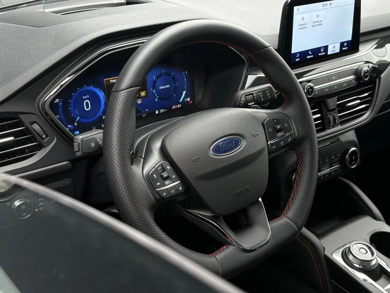 Vue intérieure centrée sur le volant noir perforé avec surpiqûres rouges du Ford Kuga 2023 et tableau de bord numérique bleu.