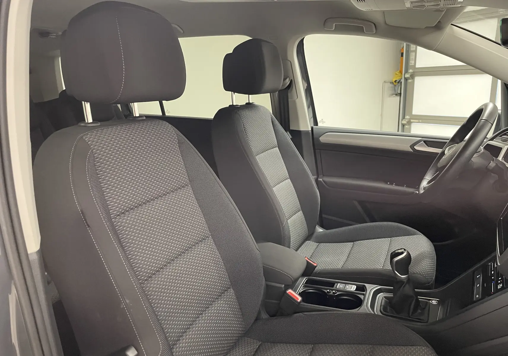 Vue intérieure côté conducteur du Volkswagen Touran 2024, sièges tissu gris avec console centrale et levier de vitesse manuel.