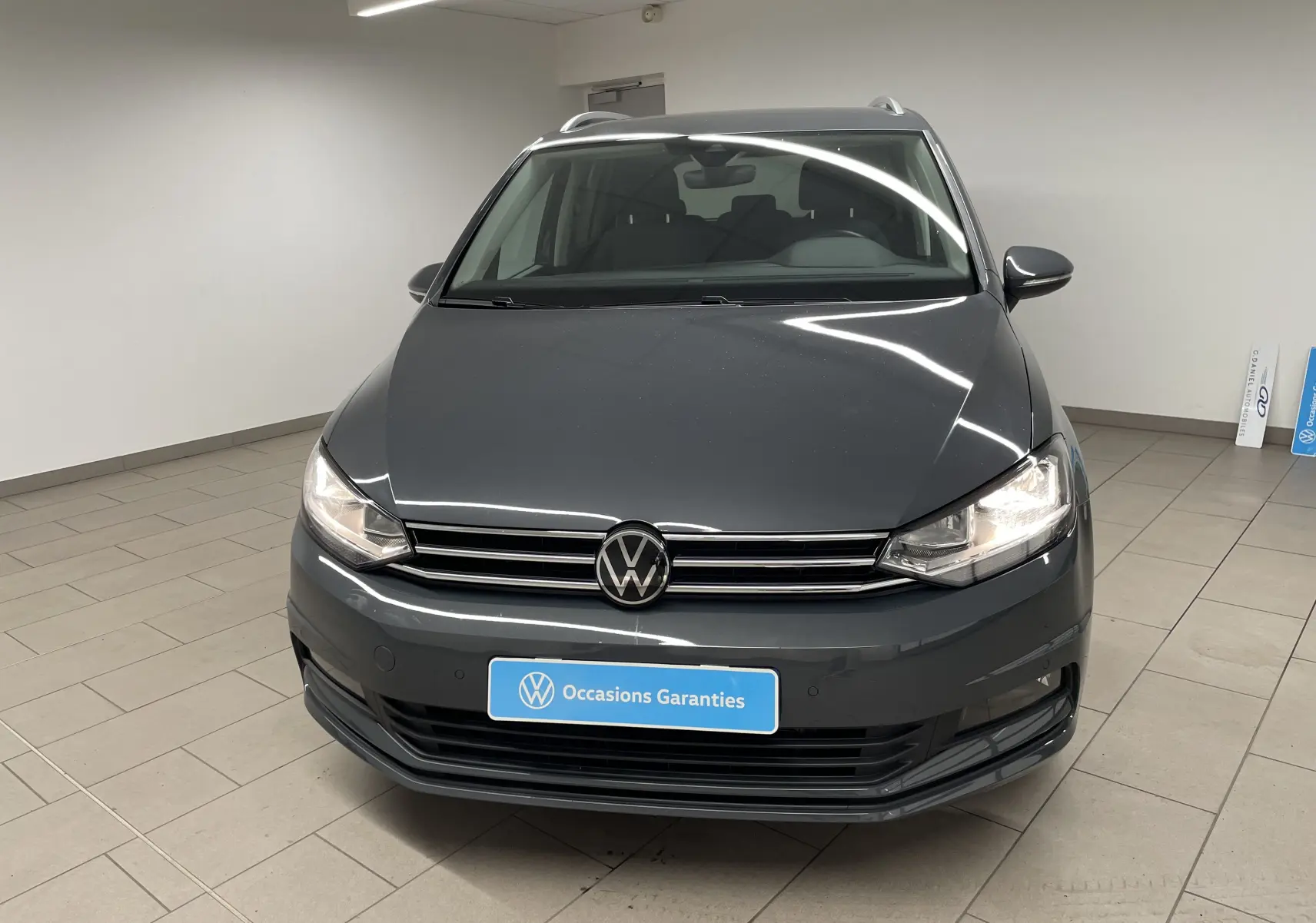 Vue de face d'un Volkswagen Touran gris dauphin 2024 avec phares allumés dans un showroom intérieur.