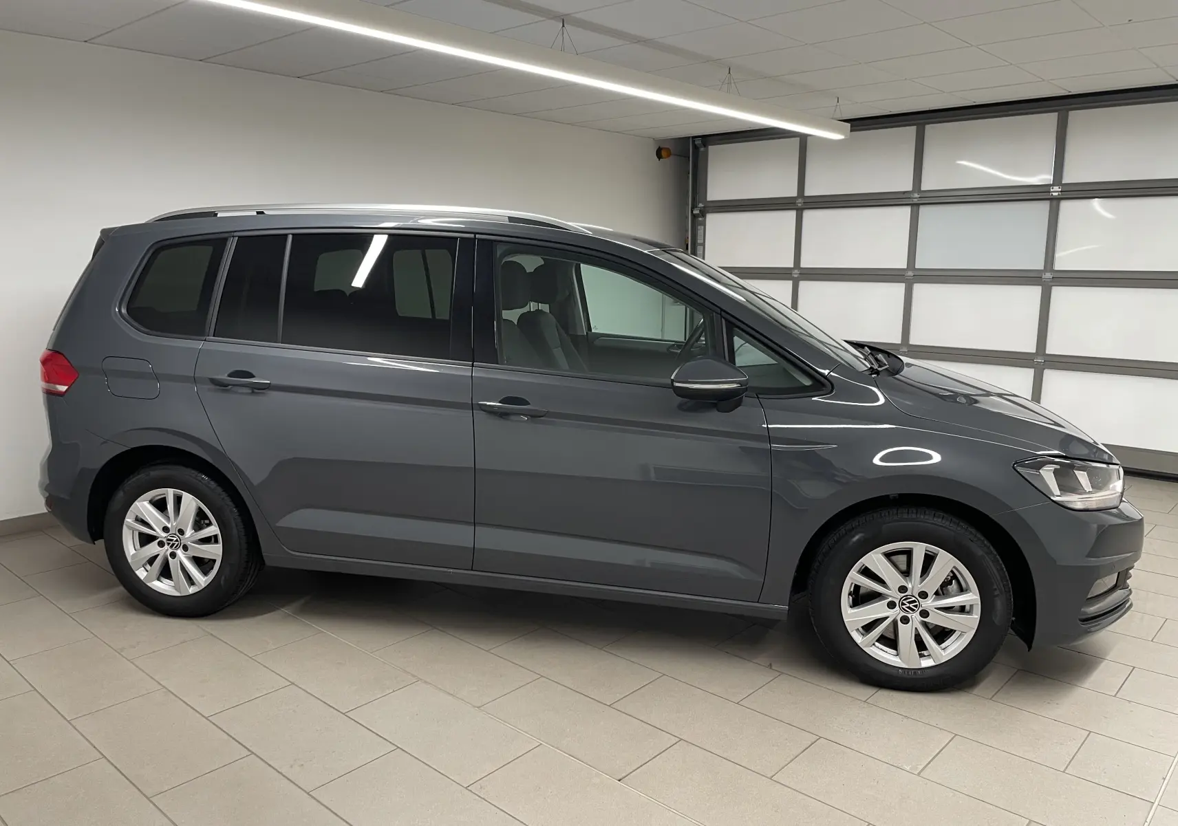Profil droit d'un Volkswagen Touran 2024 gris dauphin dans un garage, mettant en valeur sa silhouette compacte et ses jantes alliage.