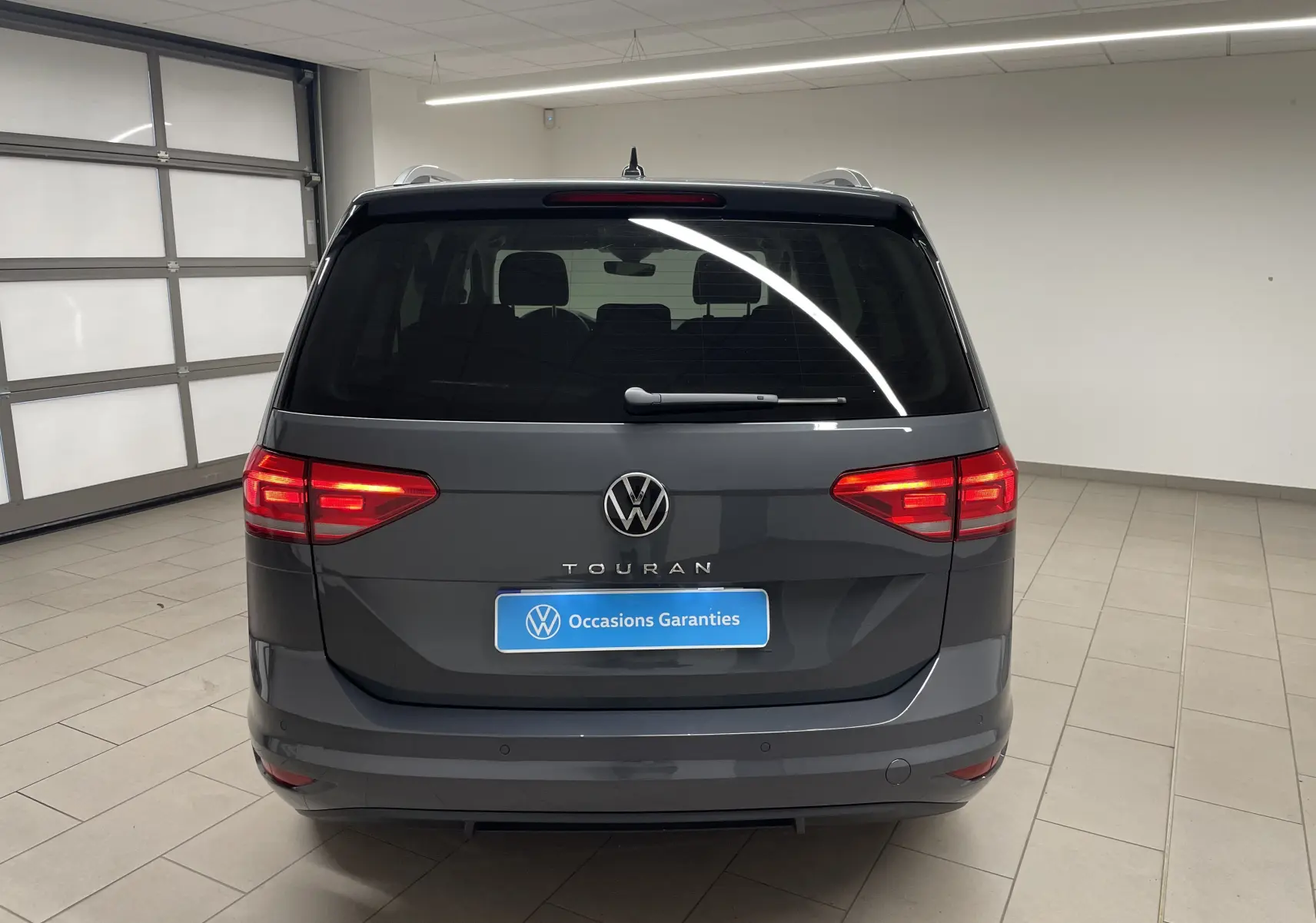 Vue arrière d'un Volkswagen Touran gris dauphin 2024 avec hayon électrique et feux arrière allumés dans un garage.