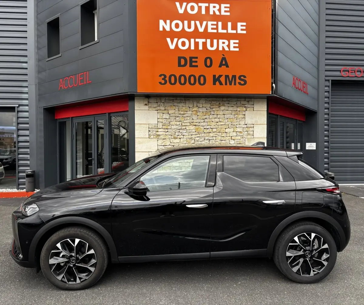 Vue profil côté gauche d'une DS3 noire 2024 avec jantes alliage et toit noir, stationnée devant un bâtiment commercial.