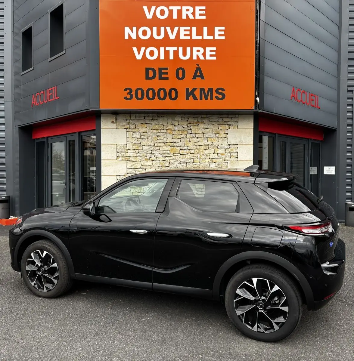 Vue latérale droite d'une DS3 noire 2024 garée devant un bâtiment avec un grand panneau orange "Votre nouvelle voiture de 0 à 30000 kms".