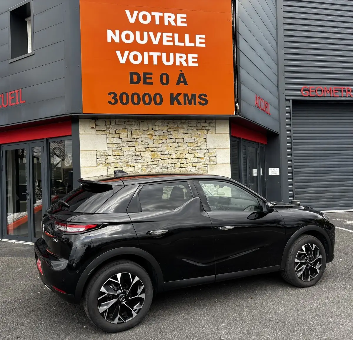 Vue 3/4 arrière droite d'une DS3 noire 2024 avec jantes alliage noires et toit noir devant un bâtiment commercial.