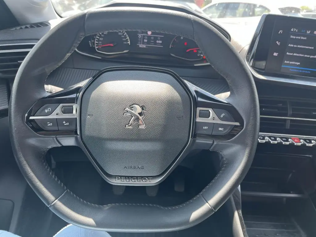 Vue rapprochée du volant noir de la Peugeot 208 2023, avec tableau de bord et écran tactile visibles en arrière-plan.
