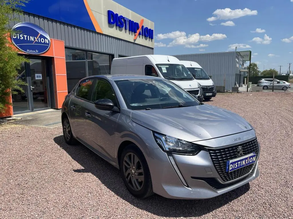 Peugeot 208 gris clair vue 3/4 avant droit, avec calandre noire distinctive, stationnée devant un garage Distinxion.