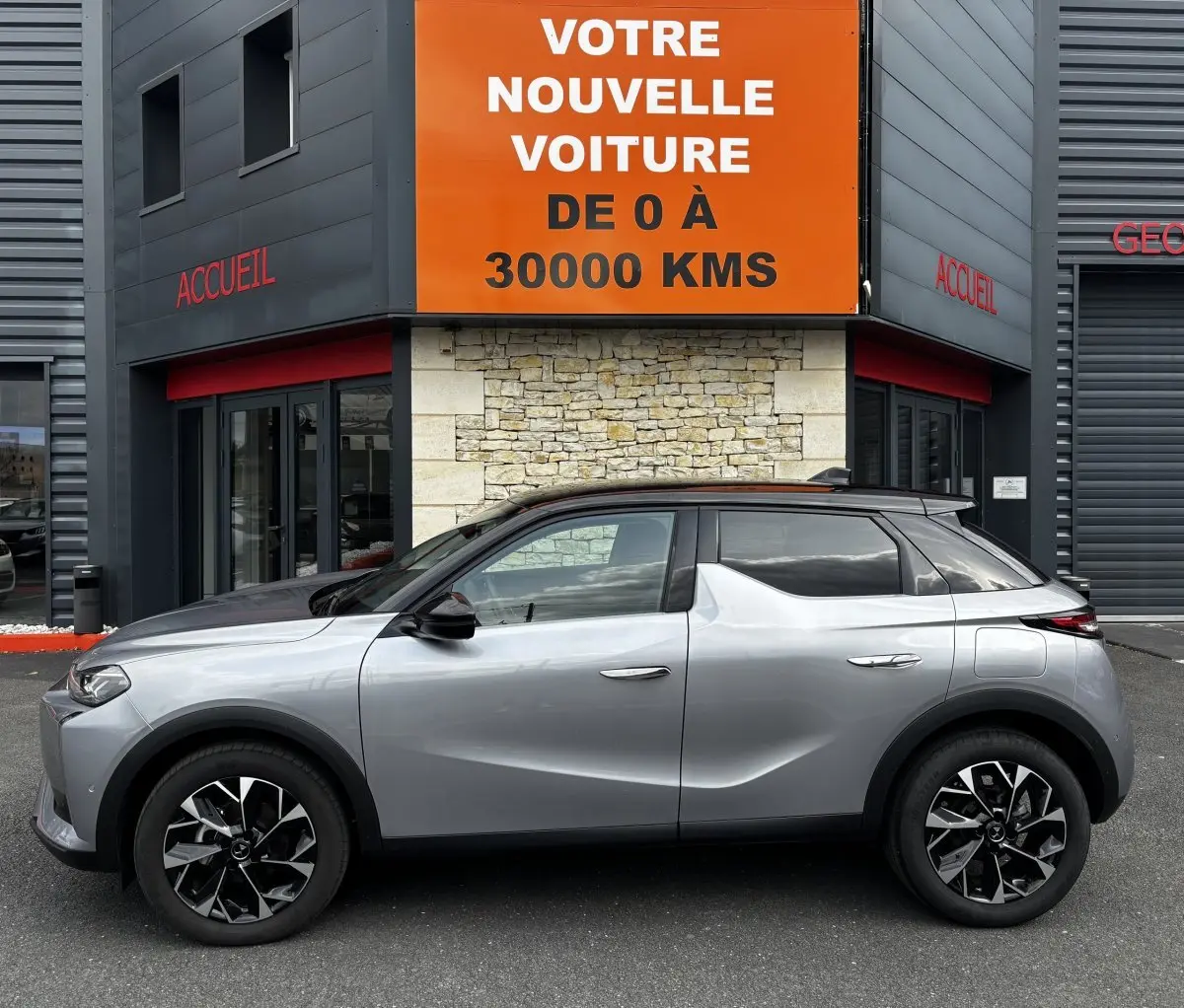 Vue latérale droite d'une DS3 2024 gris clair avec toit noir, jantes noires et détails modernes devant un bâtiment commercial.